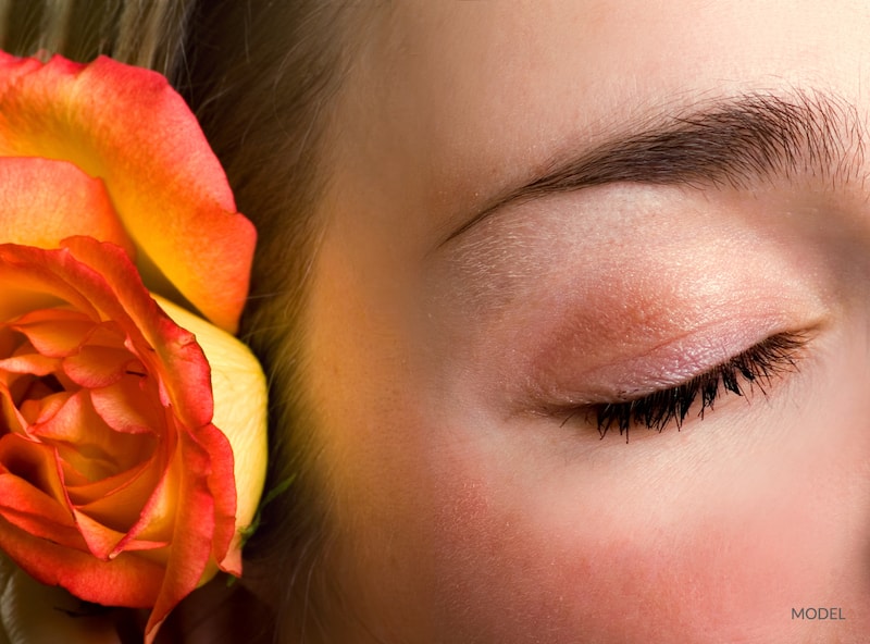 Woman with youthful, closed eye next to a orange flower. Facial rejuvenation concept.